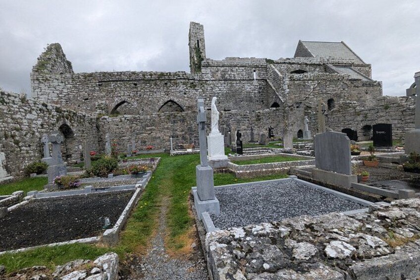 En historisk byggnad av sten, med en förfallen struktur, omgiven av en kyrkogård med gravstenar och ett centralt monument. 3