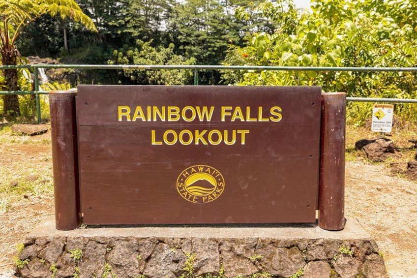 A wooden sign with 'RAINBOW FALLS LOOKOUT' and a logo, set against a natural backdrop with trees and a fence. 4