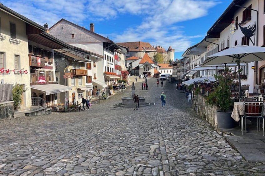 Una calle empedrada flanqueada por edificios tradicionales, una fuente y zonas de estar al aire libre. 2