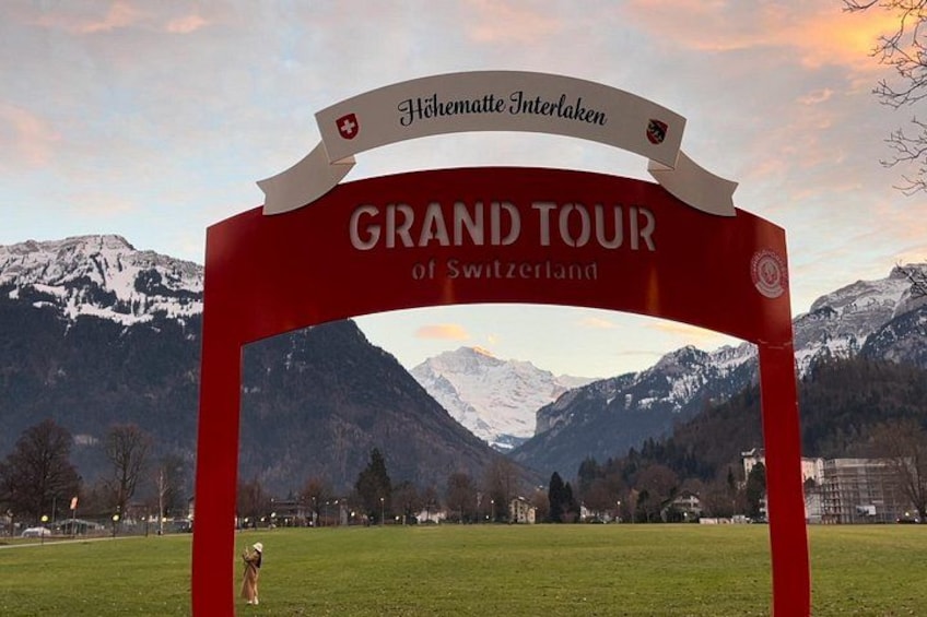 Un arco rojo con el texto 'GRAND TOUR of Switzerland' y 'Hohenschwanden Interlaken' frente a un paisaje montañoso. 3