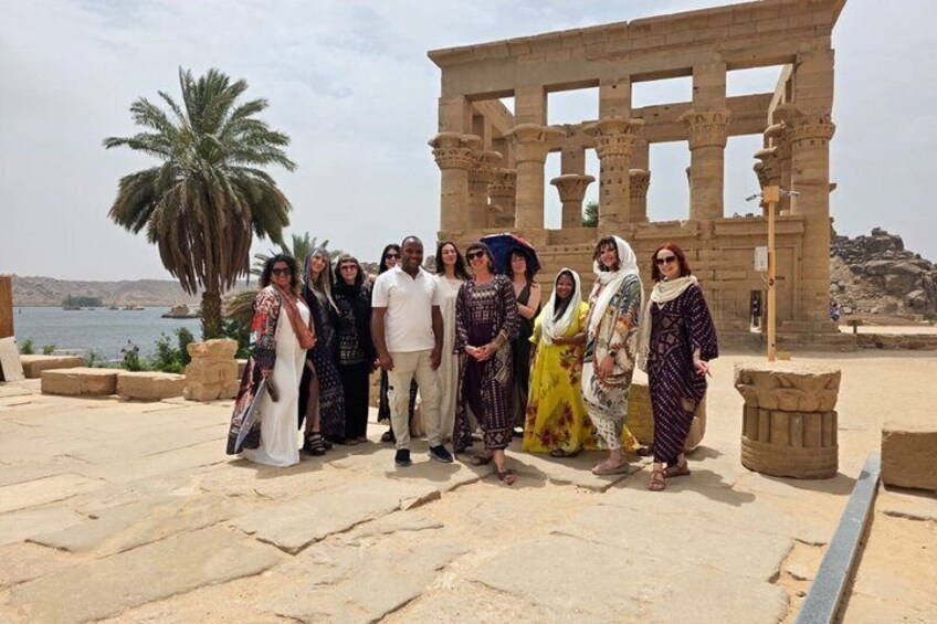 Un gruppo di persone di fronte a un antico tempio egizio, sullo sfondo palme e uno specchio d'acqua. 2