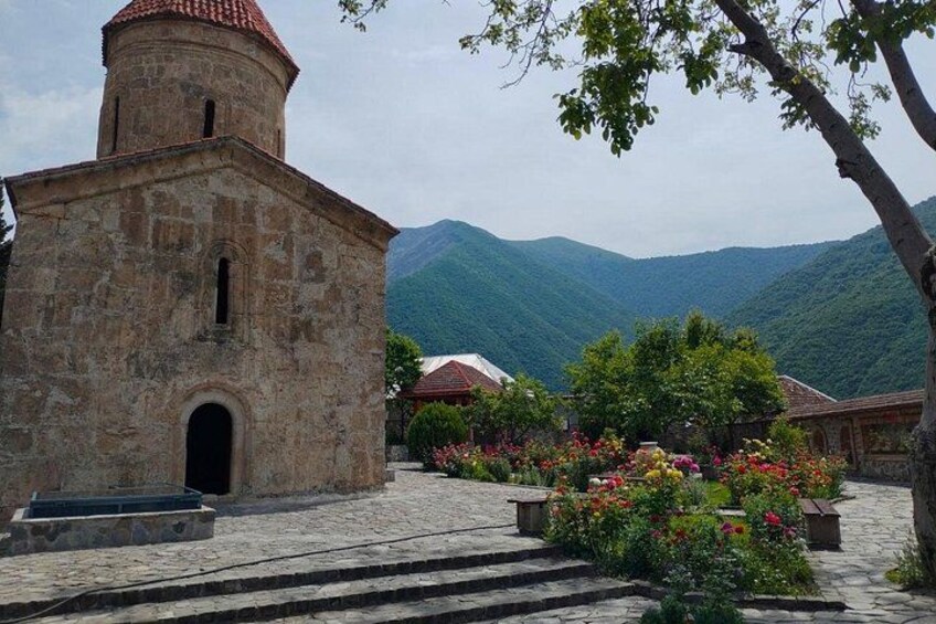 A stone church with a red-tiled roof, surrounded by a cobblestone courtyard with flowering plants. 5