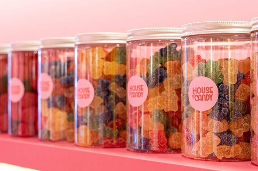 A row of clear glass jars filled with colorful gummy candies, each labeled with a pink sticker that reads "HOUSE CANDY." 2