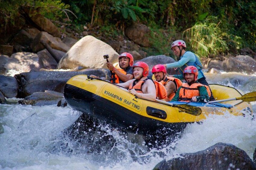 A group of people rafting on a river in a yellow raft with 'KHAMPAN RIFTING' written on it. 1