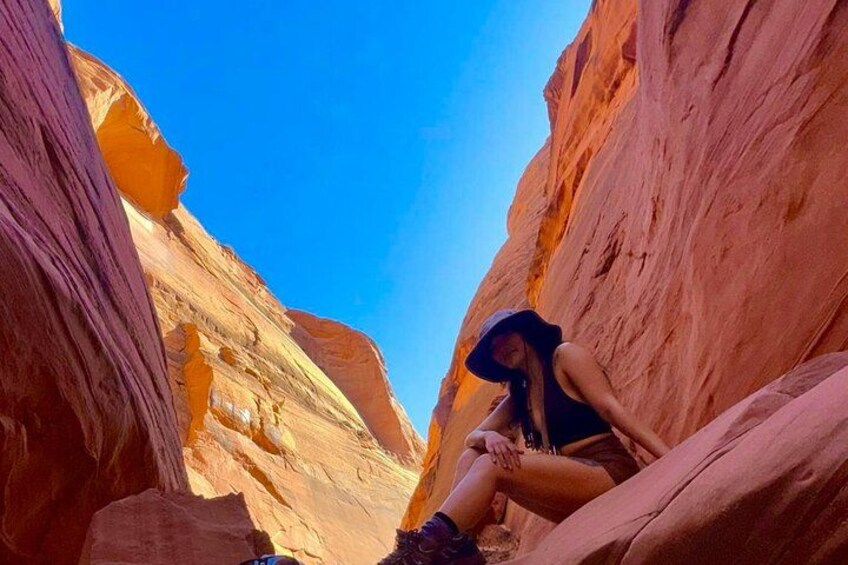 A person sitting on a rock in a narrow canyon with red rock walls and a clear blue sky. 2