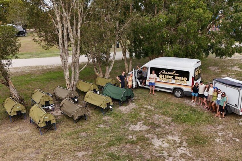 A group of people around a white bus with 'Australia Surf Bus' written on it, surrounded by trees and camping gear. 1