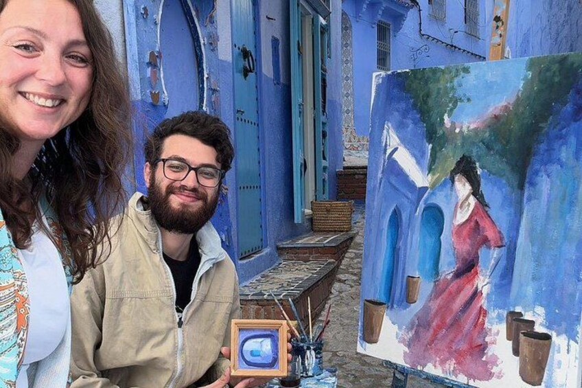 A narrow street with blue buildings and a person holding a framed painting. 1