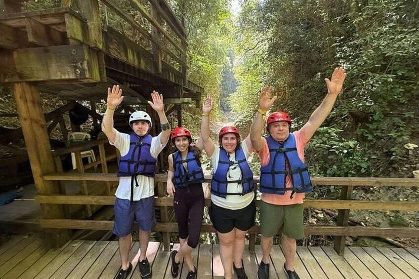 Four people on a wooden bridge with raised hands, wearing life jackets and helmets. 3