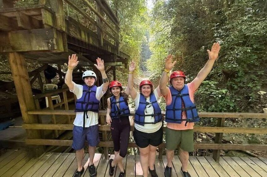 Vier Personen auf einer Holzbrücke mit erhobenen Armen, mit Rettungswesten und Helmen bekleidet. 3