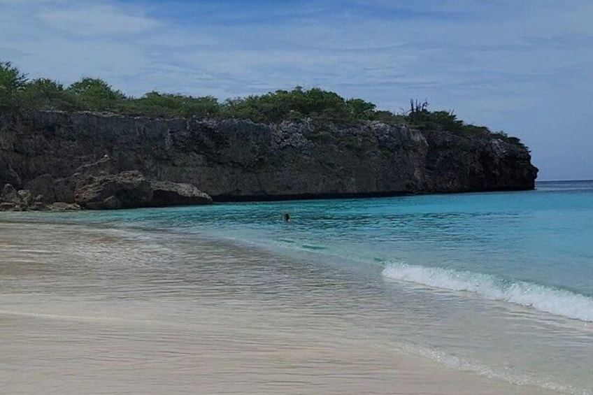 En strand med kristallklart turkosfärgat vatten, en sandstrand och en klippig brant med växtlighet. 1