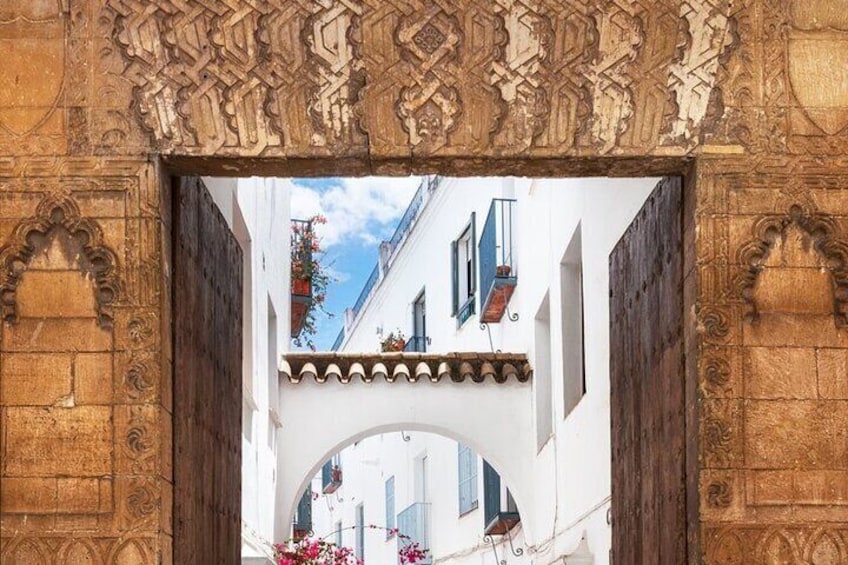 An ornate wooden archway with intricate carvings, a white building with blue shutters, and a small balcony with hanging flowers. 5