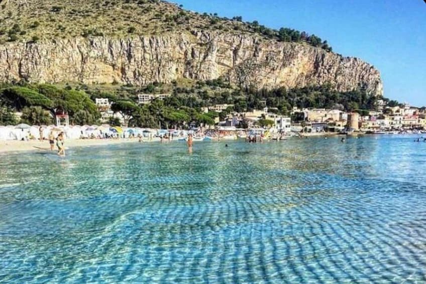 A coastal beach with clear water, people swimming, and buildings on a cliff. 2