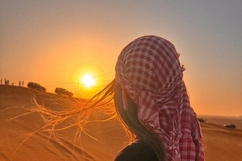Iemand in kleding met een geruit patroon loopt over zand, met een zonsondergang op de achtergrond. 1