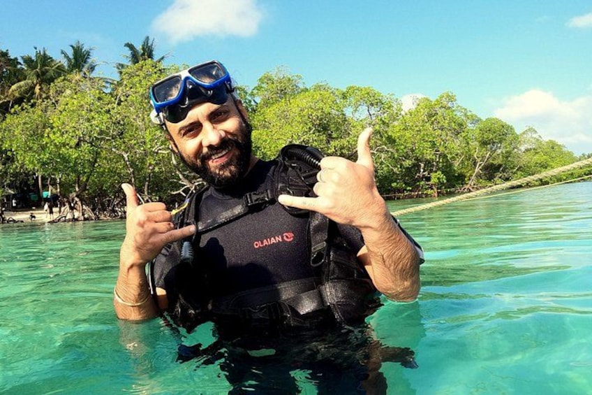 A person in a black wetsuit with a diving mask and a life vest, standing in clear turquoise water with a tropical background. 3