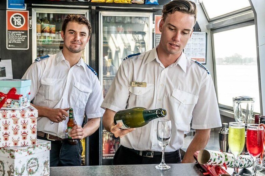 Dos hombres con uniforme sirviendo bebidas en un bar, con una caja de regalo temática navideña y un panel de menú al fondo. 3