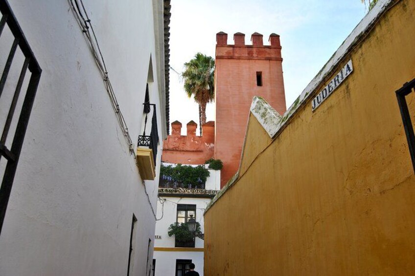 Een smalle steeg met een wit gebouw links en een geel gebouw rechts, die leidt naar een rode toren met een palmboom op de achtergrond. 5