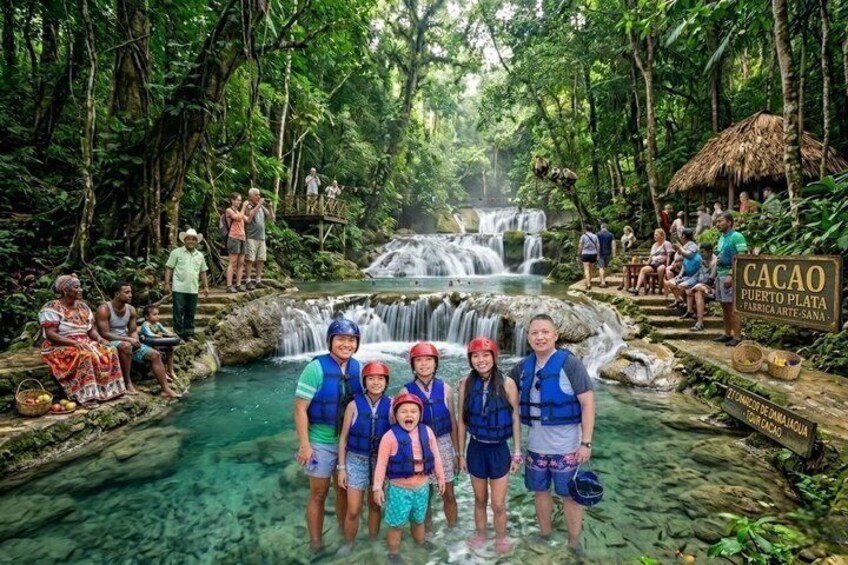 Een groep mensen in een tropische omgeving met een waterval en een bord waarop staat 'Cacao Puerto Plata Fabrica Artesana'. 1