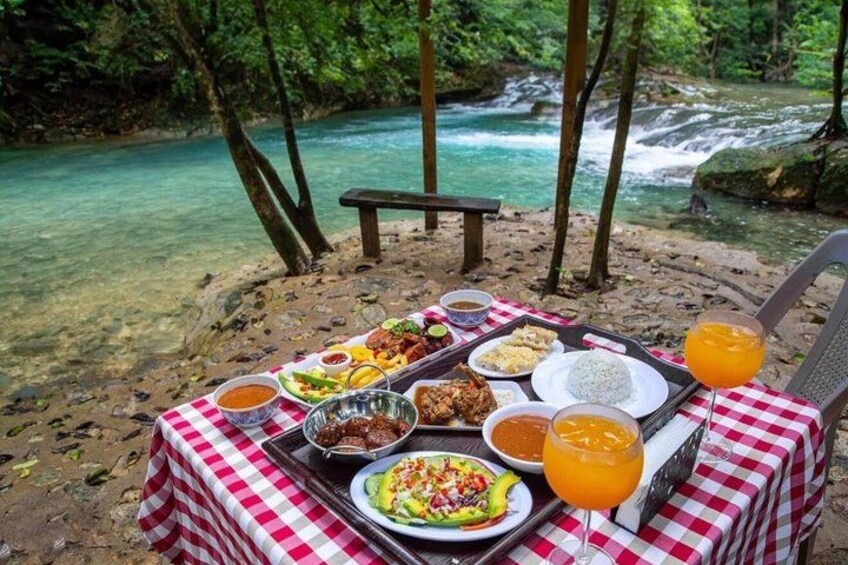 Een picknickopstelling met een geruit tafelkleed, verschillende gerechten en een glas sinaasappelsap op een zandstrand, met een rivier op de achtergrond. 5