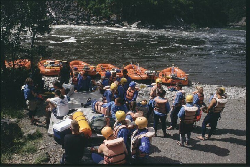 Un groupe de personnes, portant des gilets de sauvetage et des casques, s’est rassemblé près de bateaux pneumatiques sur la rive d’une rivière. 1