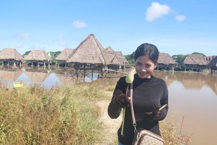A person holding a bird, standing in front of thatched-roof huts by a calm body of water. 5
