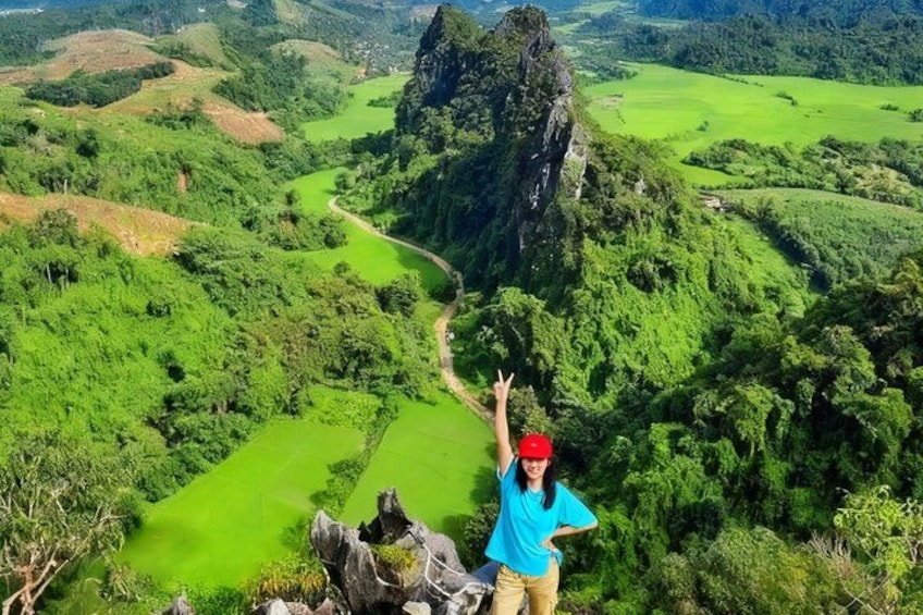 A person standing on a hill overlooking a green valley with a winding path and a prominent rock formation. 2