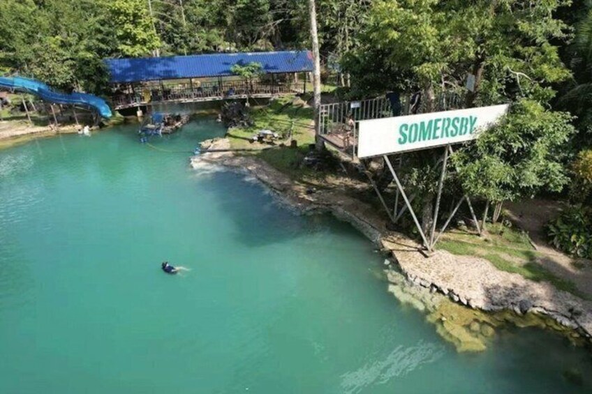 A person swimming in a clear blue pool with a sign reading 'SOMERSBY' and a blue-roofed structure in the background. 3