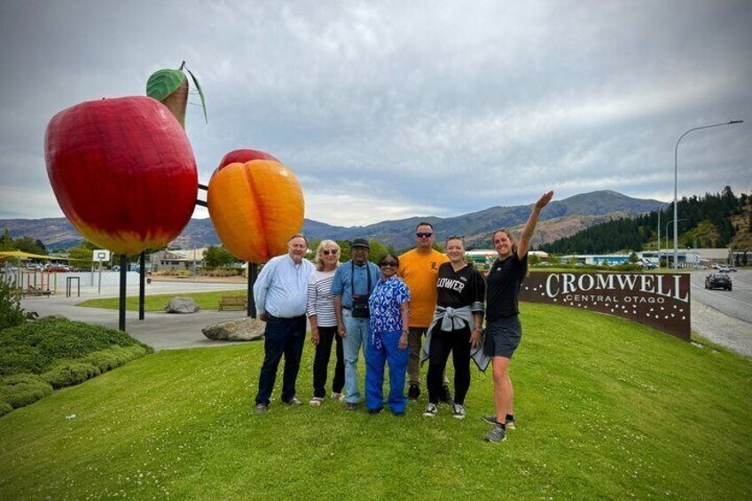 Un grupo de personas frente a un cartel que dice 'CROMWELL CENTRAL OTAGO', con una gran escultura de melocotón al fondo. 4