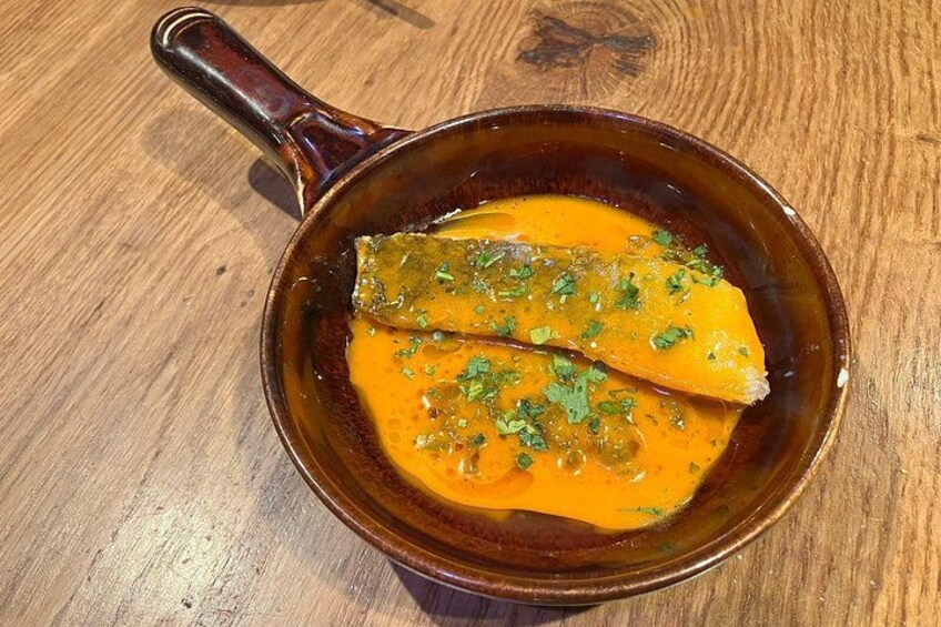 A brown ceramic bowl with a handle containing a dish with a yellow-orange sauce and green herbs, placed on a wooden surface. 3