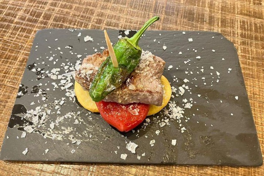 A dish with a grilled meat, a green chili, and a slice of tomato on a black slate plate. 4