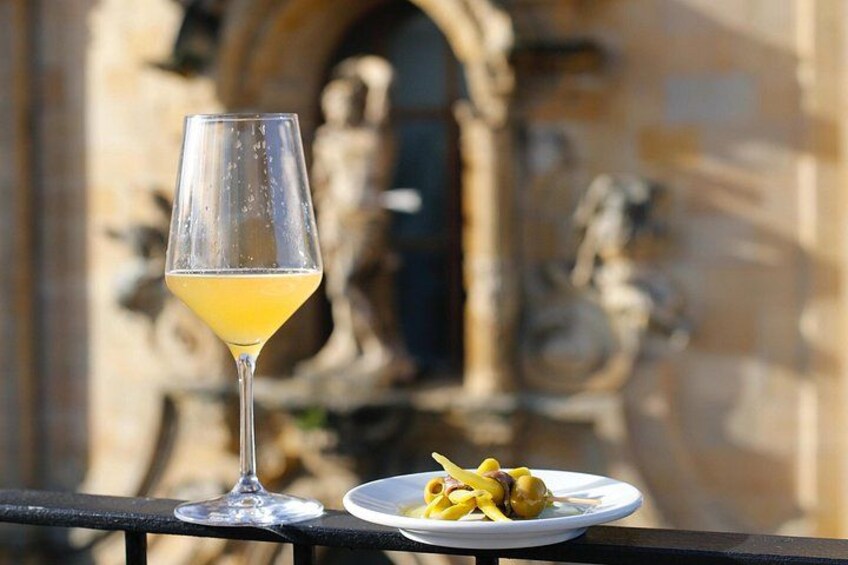 A glass of yellow drink on a balcony with a plate of food and a building with statues in the background. 1