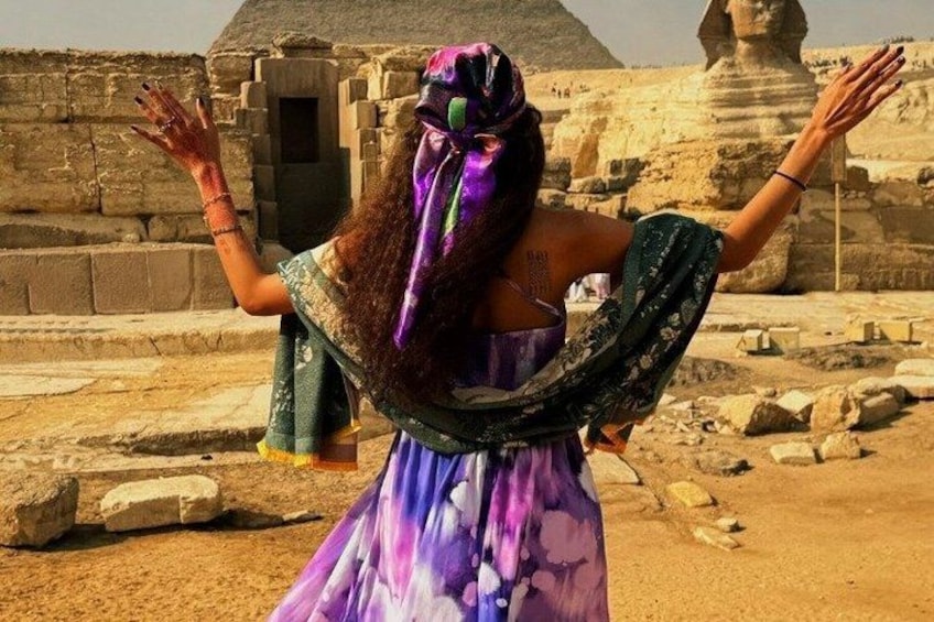 Een persoon in een kleurrijke jurk met opgetrokken armen, voor oude Egyptische ruïnes. 3