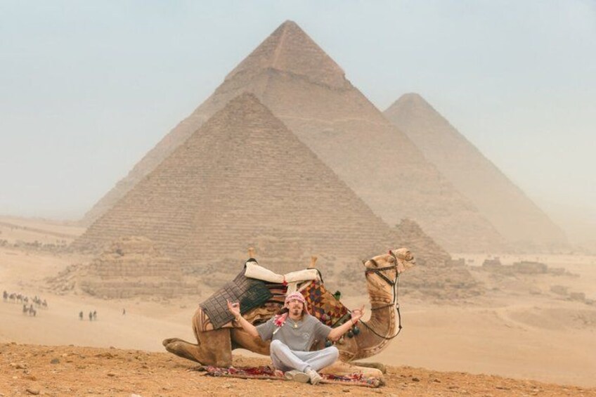 Een persoon zit op de grond, met een kameel voor de Piramide van Gizeh. 2