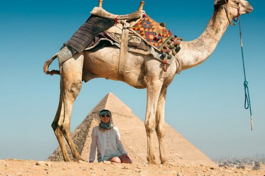 Een kameel met zadel en decoratieve deken, een persoon ernaast, en de Piramide van Gizeh op de achtergrond. 1