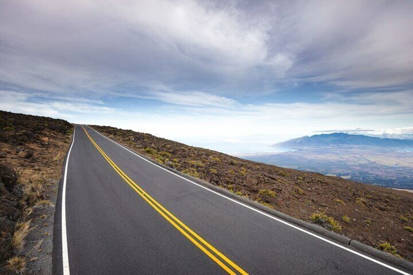 Une route à deux voies, délimitée par des lignes jaunes, bordée d’un paysage rocheux et d’une végétation clairsemée, qui mène vers des montagnes lointaines sous un ciel nuageux. 5