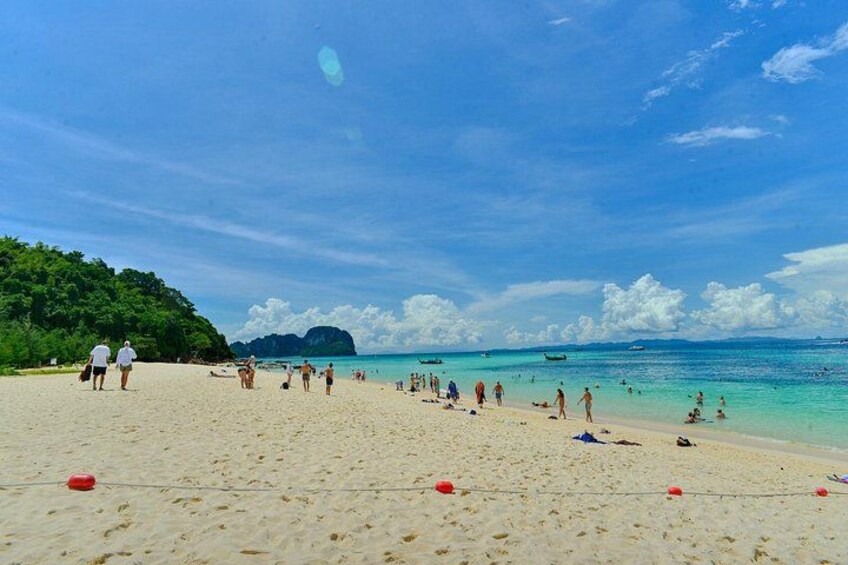 A beach with people walking and swimming, clear blue water, and a distant mountain. 4