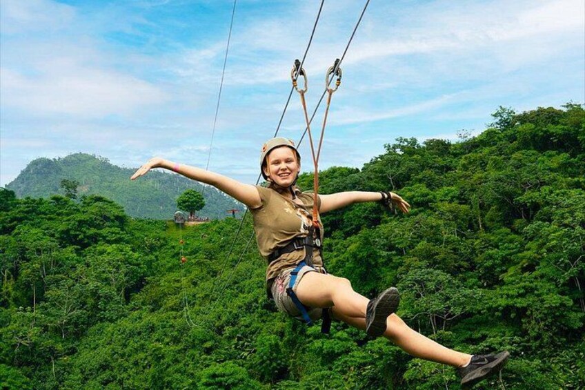 A person zip-lining through a lush green forest with dense foliage and a distant hill in the background. 1