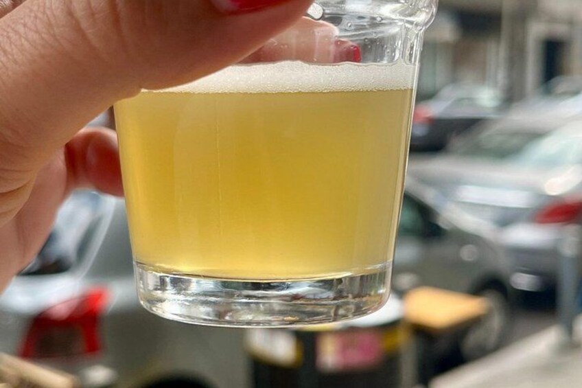 A hand holding a glass of beer with a frothy head, set against a blurred urban background. 1