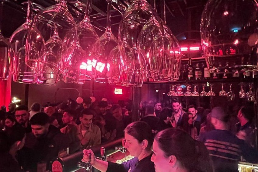 A crowded bar with people socializing, a bartender, and hanging wine glasses. 5