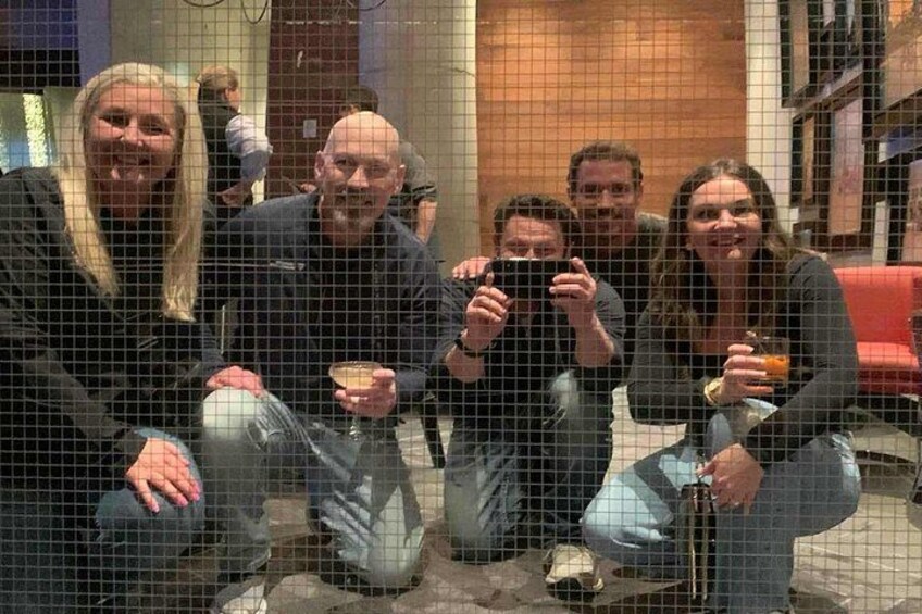 A group of people sitting on the floor, holding drinks, in a room with a red couch and a wooden door in the background. 2