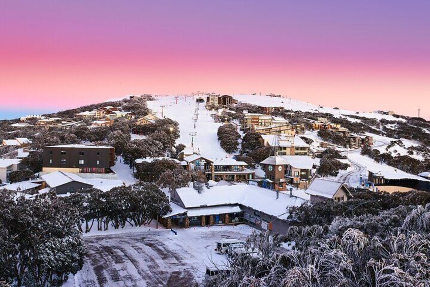 A snow-covered village with buildings and trees, under a pink sky. 1