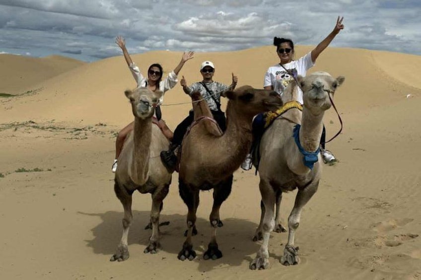 Three people riding camels in a desert with sand dunes in the background. 3