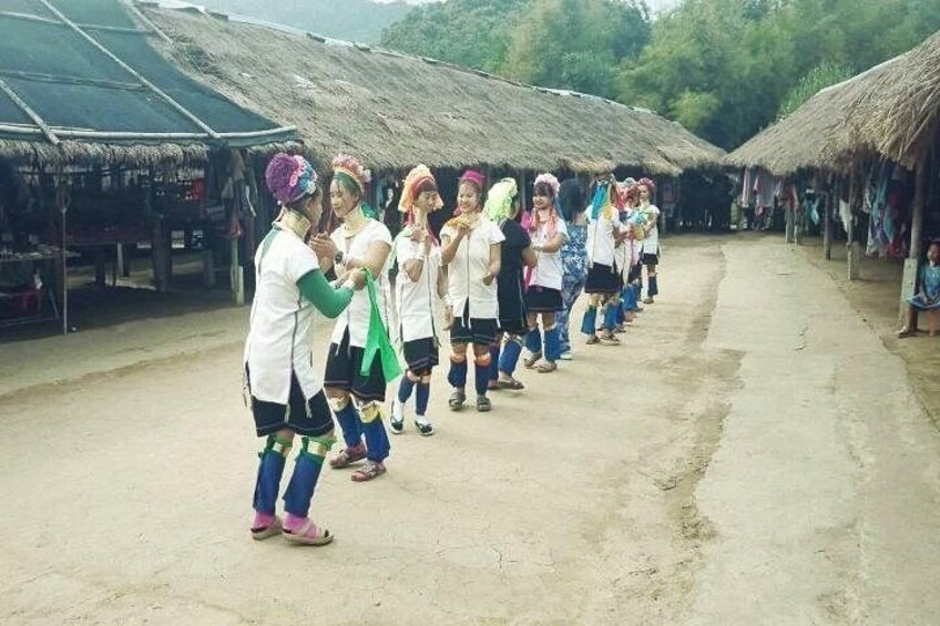 A group of people in traditional attire lined up on a dirt path between thatched-roof structures. 5