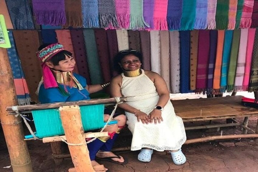 Two people sitting on a bench under a colorful woven roof, with a variety of textiles hanging in the background. 2
