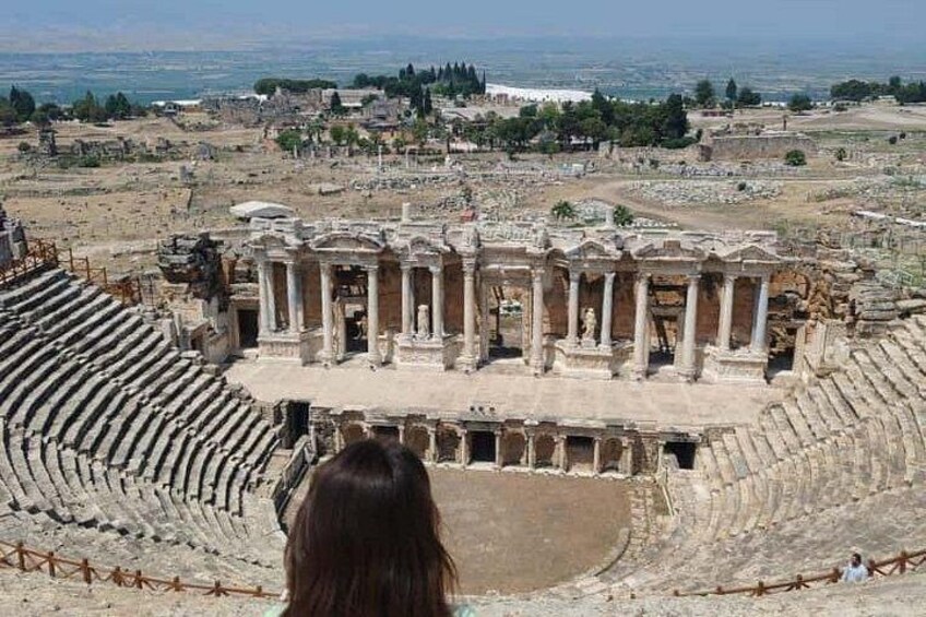 Un antiguo anfiteatro con columnas y escenario, rodeado de ruinas y un paisaje urbano distante. 4