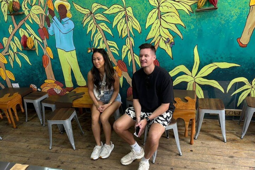 Two people sitting on stools in a room with a mural of a man harvesting cocoa pods. 3