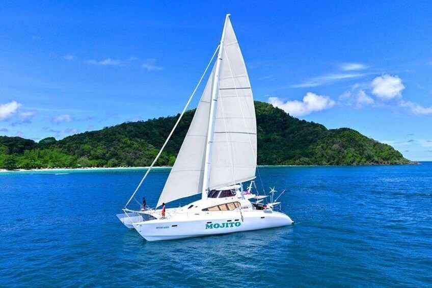 A white sailboat with a white sail on a calm sea, surrounded by clear blue water and a green island in the background. 1