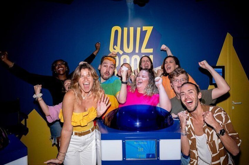A group of people celebrating in front of a blue and yellow backdrop with the word 'QUIZ' visible. 1