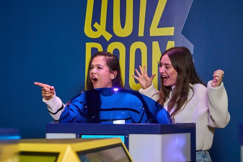 Two people at a quiz booth with a large 'QUIZ ROOM' sign in the background. 2
