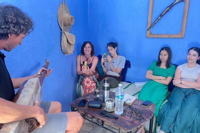 A group of people sitting around a table with a guitar player, a blue wall, and a woven basket hanging on the wall. 3