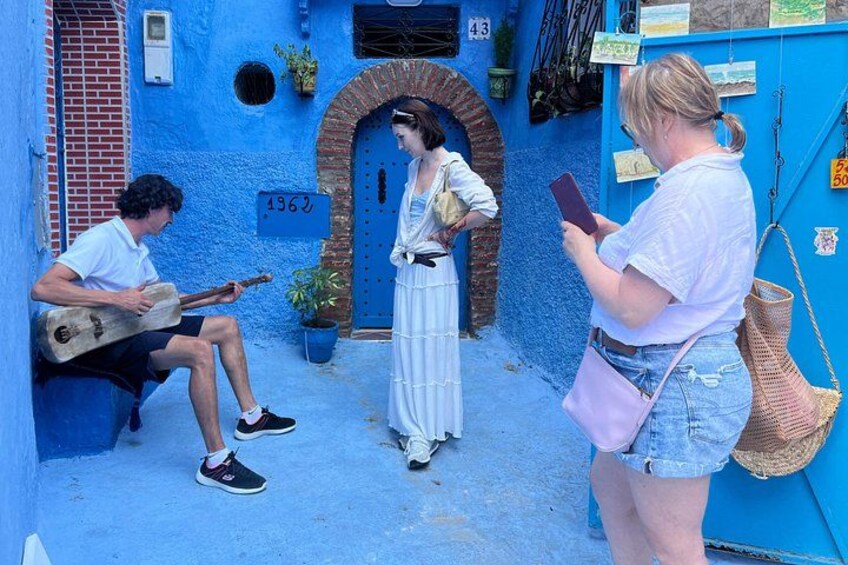 A narrow alley with a blue wall, a person playing a guitar, and another person taking a photo. 5